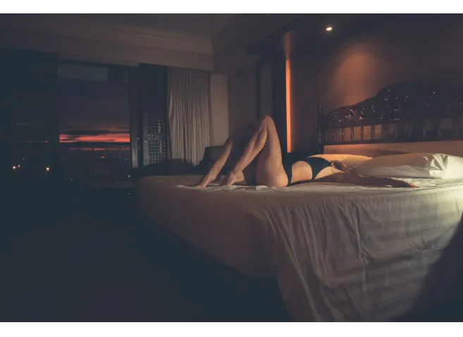 A sexy young woman in a swimsuit is lying on a hotel room bed at sunset